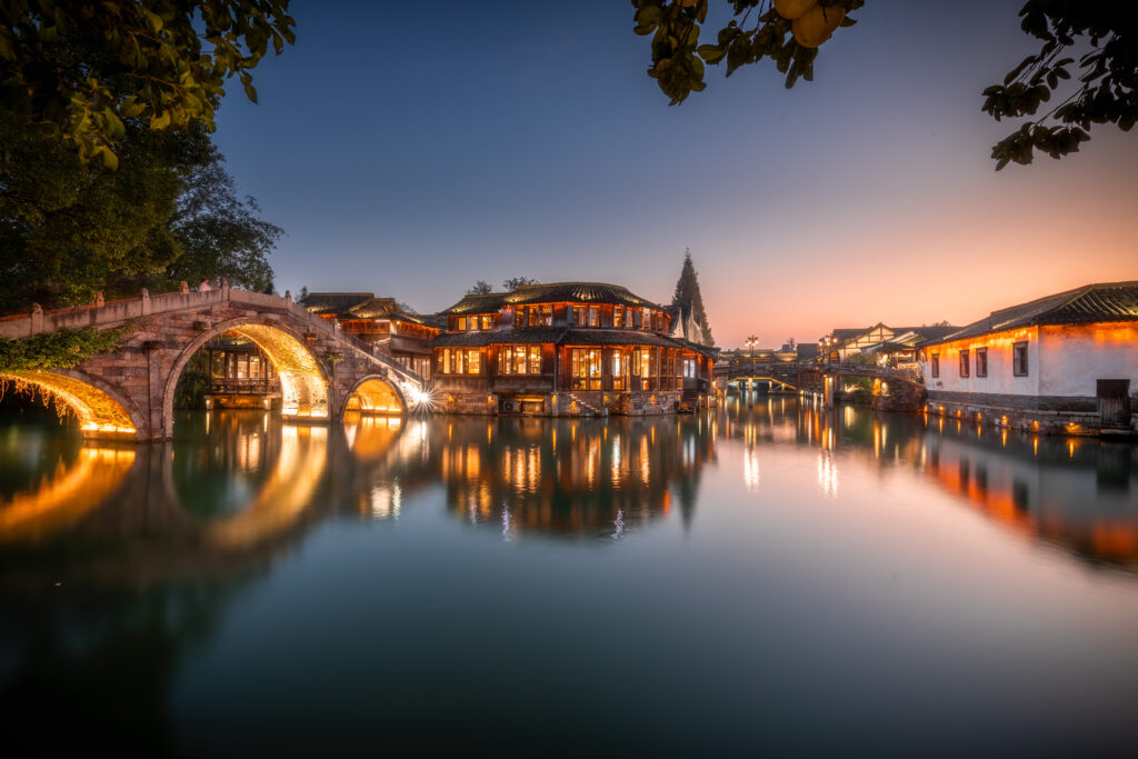 An illuminated stone bridge with round arches and traditional houses is reflected in the water of a canal at sunset; the sky shows color gradients from blue to orange.