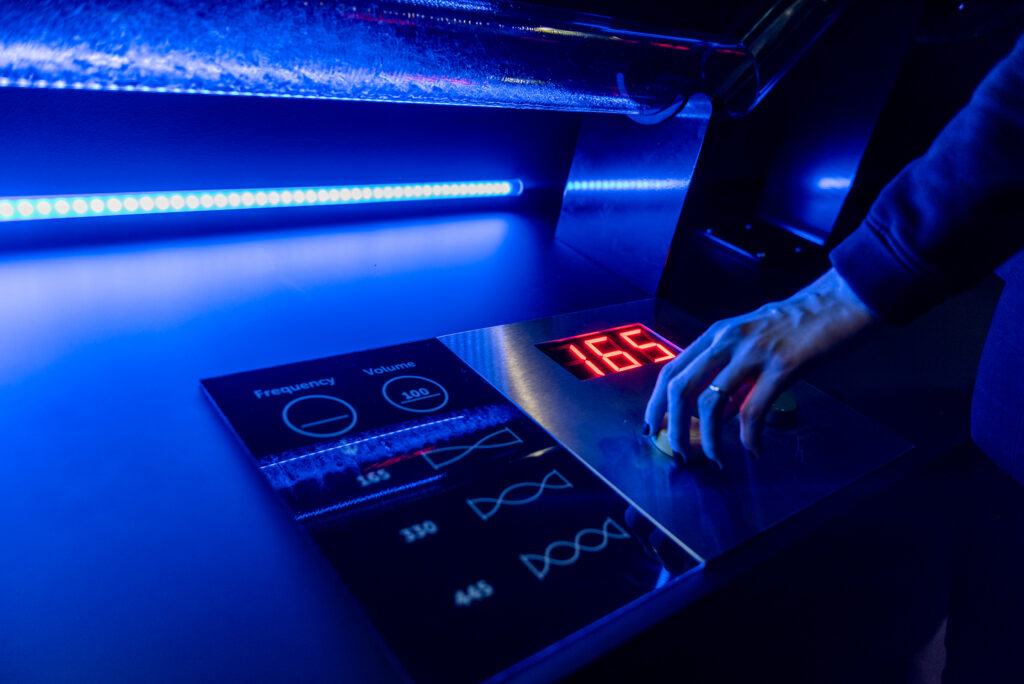 A handheld control panel with blue backlighting displays a digital screen with the number 165 as well as graphic symbols for frequency and volume, while a hand presses a button.