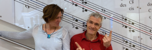 Eine Frau im hellblauen Oberteil mit blauem Anhänger und ein Mann im roten Hemd sitzen vor einer Wand mit großen Notenlinien und Musiknoten.