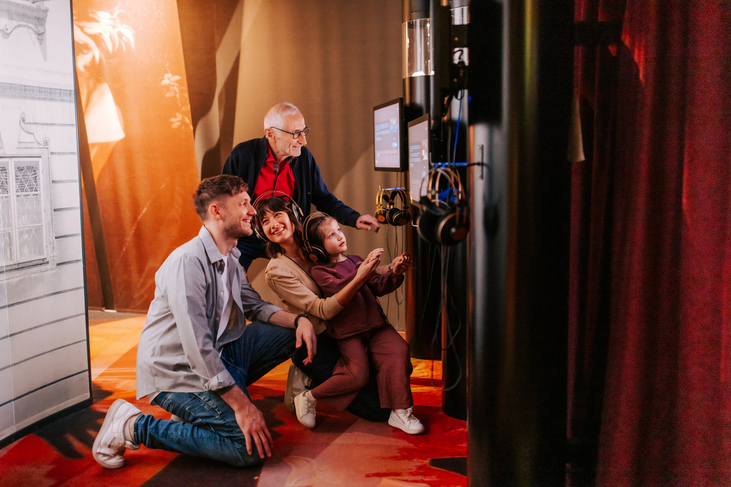 Four people, including a child, sit and stand in front of interactive screens with headphones connected while using the exhibition; red curtains and orange flooring can be seen in the background.
