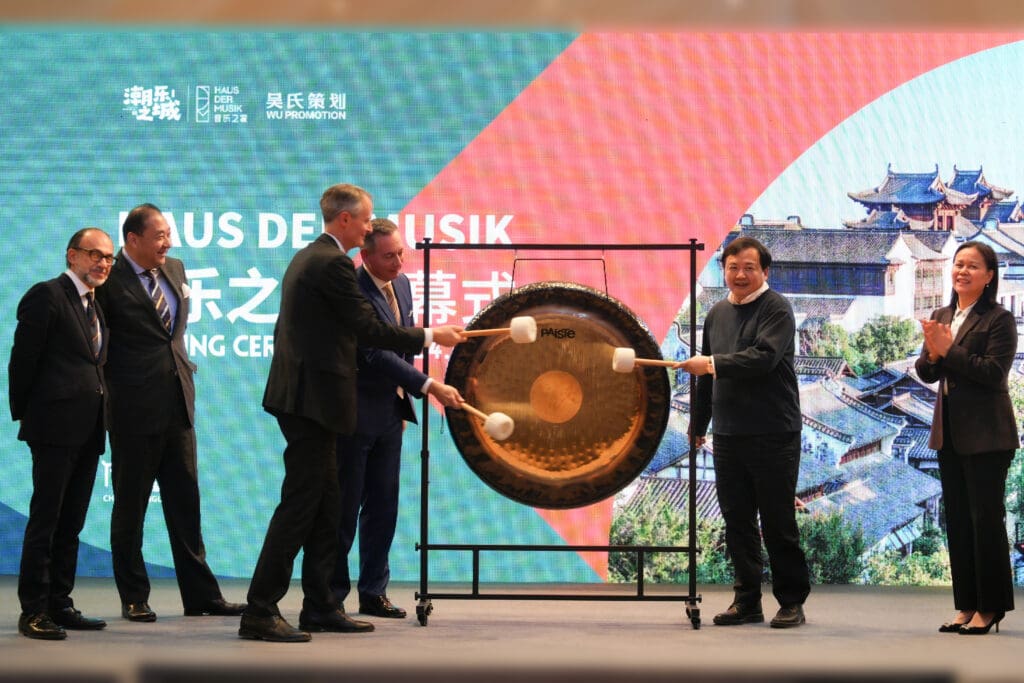 Several people in suits and business attire stand on a stage in front of a screen with the words "House of Music" in German, English and Chinese, while three men strike a large gong together with mallets.