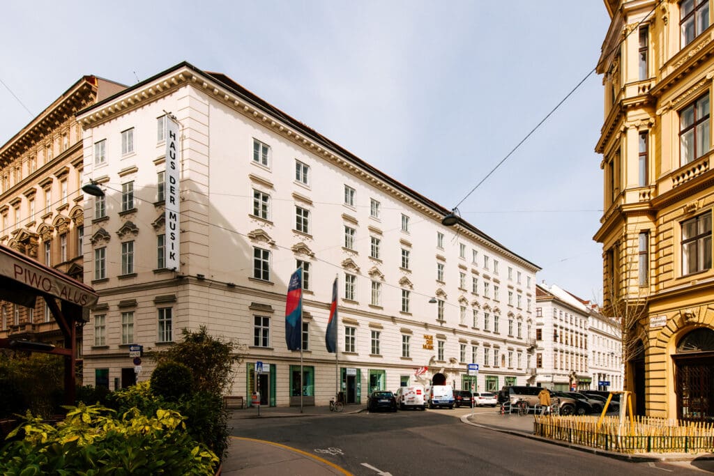 Ein großes helles Eckgebäude mit der Aufschrift „Haus der Musik“ und mehreren wehenden Fahnen steht an einer Straßenecke, davor parken Autos und Fahrräder, umgeben von historischen Nachbarhäusern.