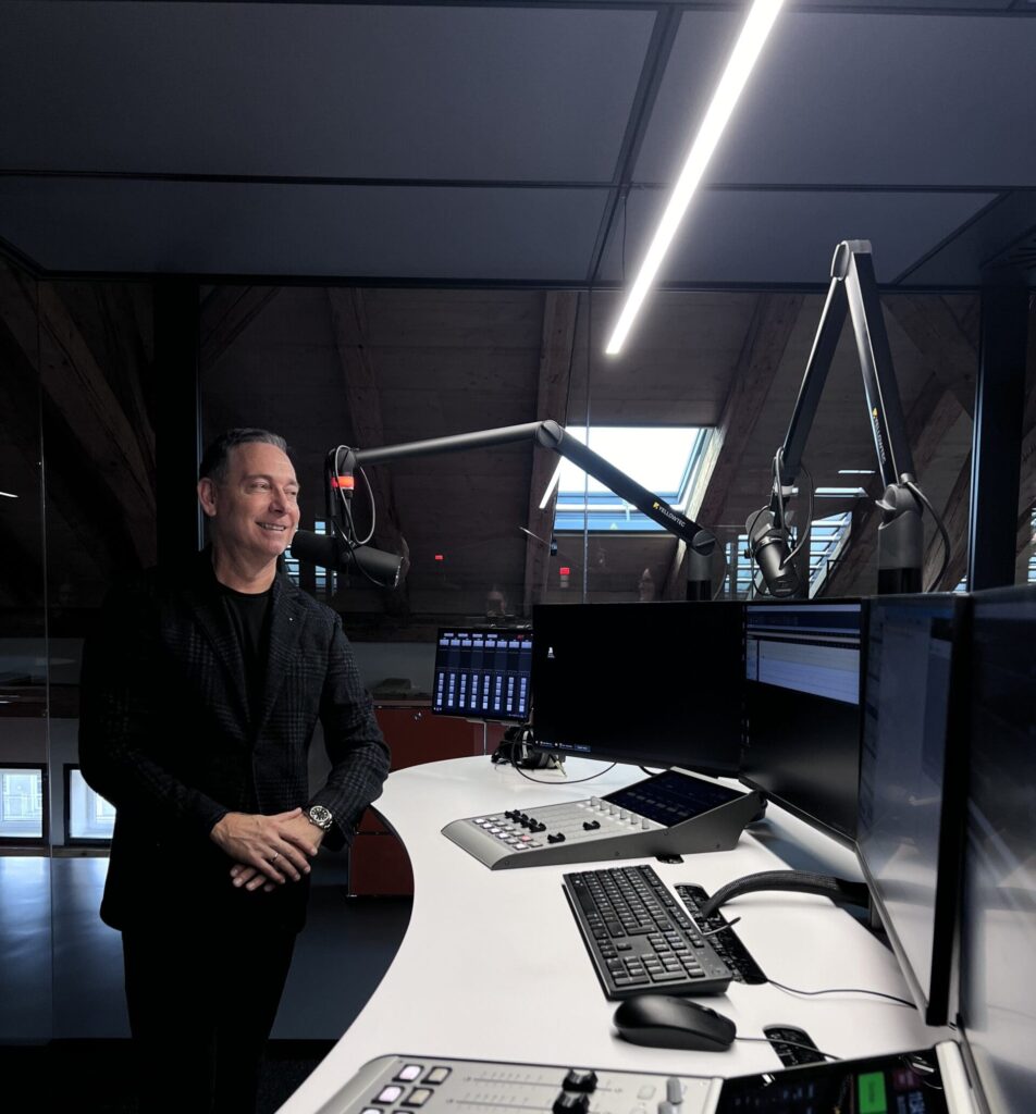 A man stands smiling in a modern radio studio in front of a table with mixing console, keyboard, monitors and microphone arms; in the background a glass wall with a skylight can be seen.