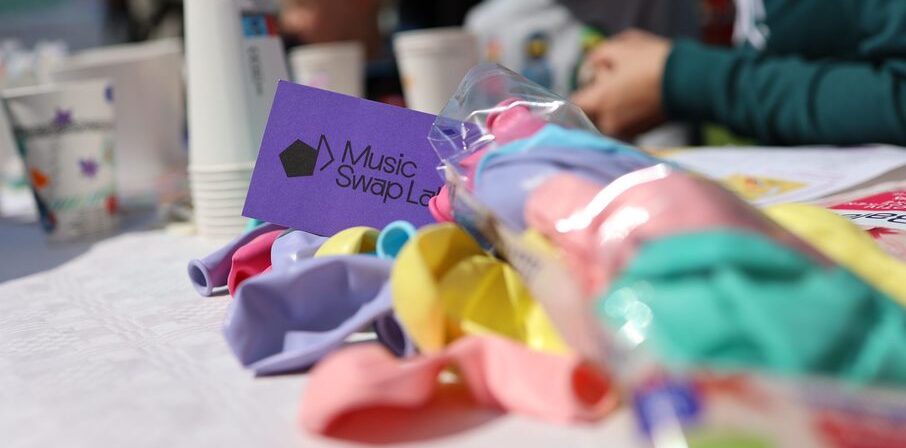 Auf einem Tisch liegen bunte Luftballons und eine violette Karte mit der Aufschrift „Music Swap Lab“, im Hintergrund sind Pappbecher und unscharf Personen zu erkennen.