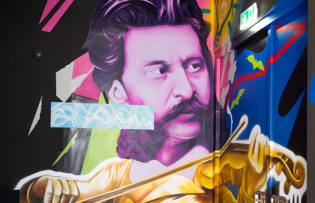 Strauss Mural von Max Rocks - Buntes Wandgemälde mit dem Porträt eines Mannes in Violett- und Pinktönen, der eine Geige mit goldgelbem Bogen spielt, umgeben von abstrakten Formen und kräftigen Farben.
